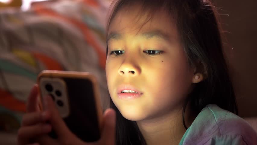 Asian girl is looking at her mobile phone in low light and the blue light from the phone is harmful to her eyes. Cute girl is looking at smartphone on sofa late at night, screen addiction in children.