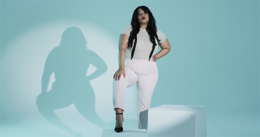 Woman entering mint-green studio adjusting suspenders smiling and modeling outfit on white pedestal. Fashion, portrait, studio, minimalist, elegance, confidence, femininity