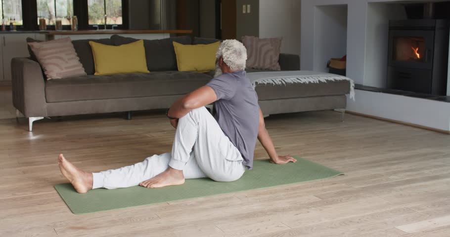 Senior African American man in sweatpants twisting on yoga mat at home holding twist, repeating. Wellness, flexibility, mindfulness, home fitness, meditation, relaxation, active aging