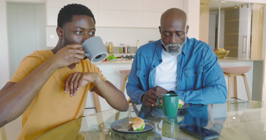 African American father and son sipping from mugs slicing sandwiches in modern kitchen chatting. Family, mentorship, communication, bonding, cozy, contemporary, lifestyle