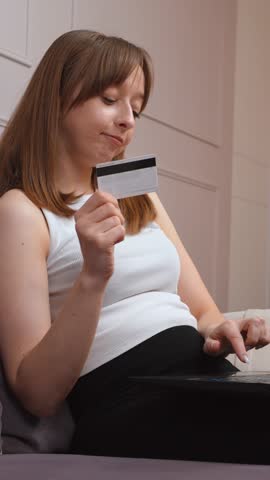 Comfortable casual woman sitting on sofa at home, holding credit card and typing payment information on laptop, enjoying convenient online shopping experience vertical video