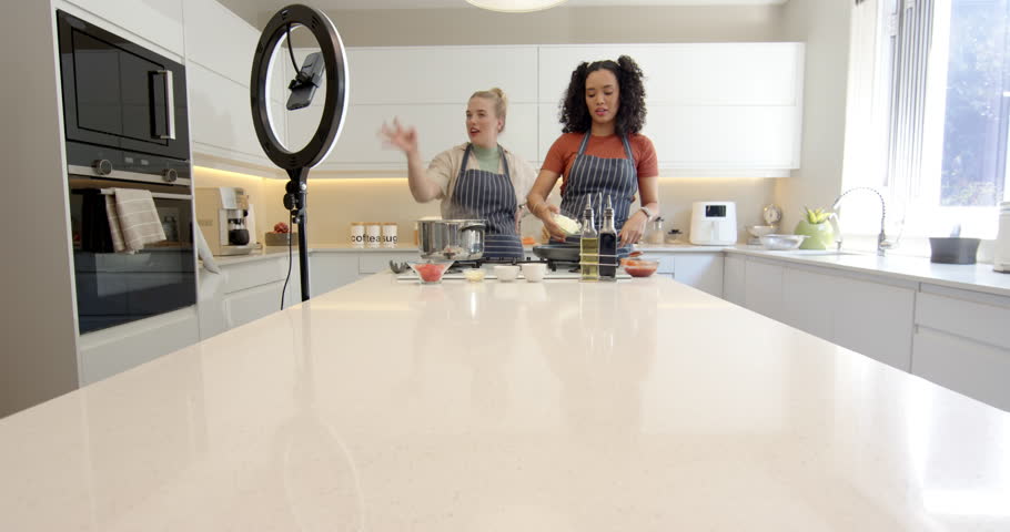 Diverse female chefs greeting camera, pointing spices, stirring skillet in kitchen for cooking demo. Cooking, culinary, modern, vibrant, teamwork, collaboration, lifestyle