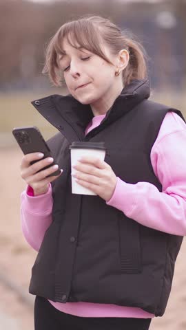 Young woman enjoys a coffee while checking her phone during a leisurely stroll through a peaceful park, embracing the tranquility of nature and the convenience of modern technology vertical video
