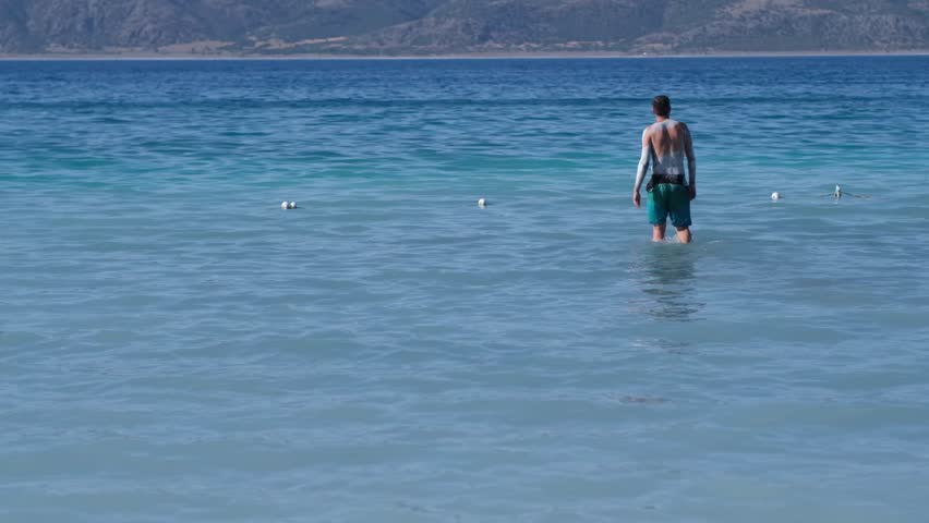 A person wades into clear, calm water along the shore, exploring the healing properties of natural mud with stunning mountain views in the background.