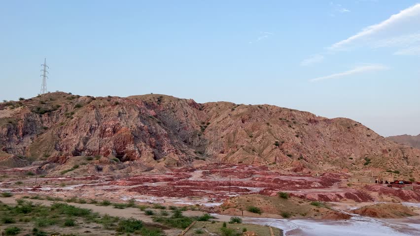 A vast, rocky hill landscape stretches under a clear blue sky, with a solitary power transmission tower standing tall on the horizon, symbolizing human presence in nature.