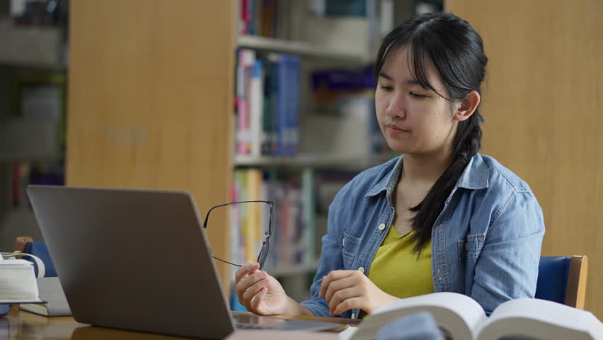 A young, tired Asian female student rubs her eyes due to strain while studying late on her laptop in a library, conveying concepts of stress, fatigue, overwork, and eye strain. - Powered by Shutterstock - Get 15% off with code: PIKWIZARD15
