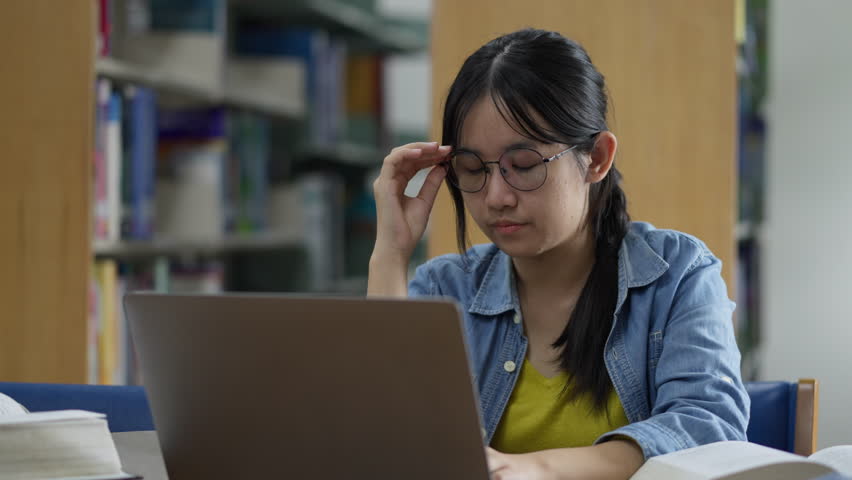A young, tired Asian female student rubs her eyes due to strain while studying late on her laptop in a library, conveying concepts of stress, fatigue, overwork, and eye strain. - Powered by Shutterstock - Get 15% off with code: PIKWIZARD15