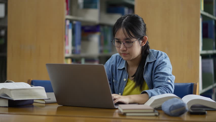 A young, tired Asian female student rubs her eyes due to strain while studying late on her laptop in a library, conveying concepts of stress, fatigue, overwork, and eye strain.