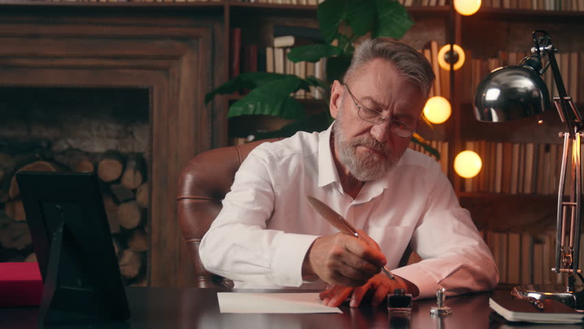 Old Caucasian European pensioner writing using fountain pen dipping ink into inkwell elder man male businessman text paper business office health medicine indoor paperwork professor author writer poet
