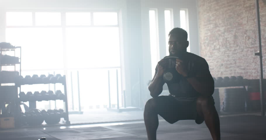 African American male gripping kettlebell and squatting, pushing upward and repeating in gym. Fitness, strength, workout, training, athleticism, determination, modern
