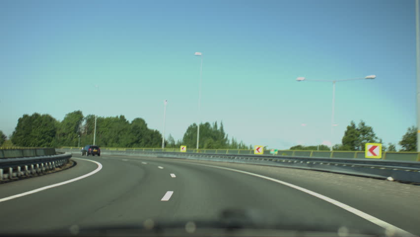 Point-of-view footage from inside a car driving along a smooth, curved highway surrounded by lush greenery. The scene captures the sense of travel, movement, and freedom on a clear summer day.