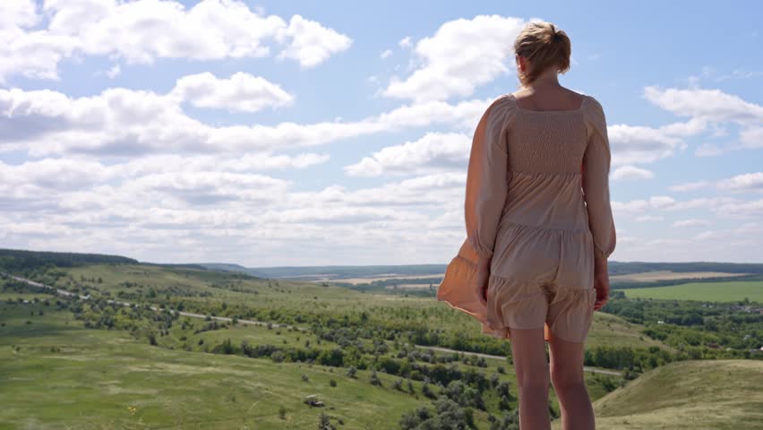 Blonde woman in a light dress stands on a hilltop, gazing at the vast green valley below, her dress swaying gently in the wind under a bright blue sky dotted with white clouds
