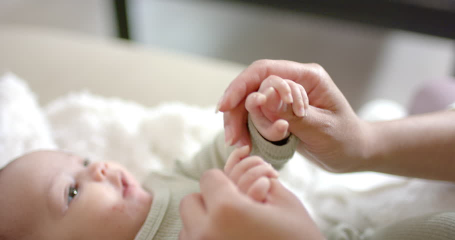 Mother lifting infant wrist, uncurling fist and massaging fingers on blanket on bed, soothing baby. Care, bonding, tenderness, intimacy, nurturing, childhood, health