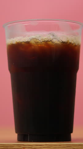 4K footage vertical video Front view SLO MO CU, iced coffee in clear plastic cup milk is poured in beautiful swirling patterns against pink background, ideal for beverage cafe or refreshment.