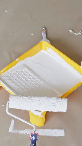 Vertical close-up showing two white paint rollers and a yellow tray filled with white paint, highlighting a renovation process involving wall or ceiling painting preparation.