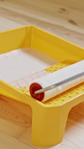 Vertical Close-up of a paint roller being evenly coated with white paint in a bright yellow tray, placed on a wooden floor, showing the preparation phase of an indoor renovation project.