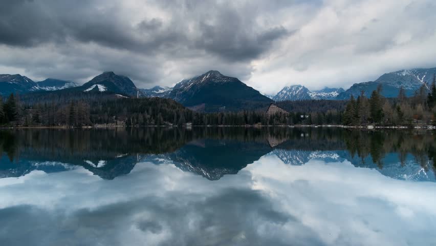 4K wide shot of snowy Tatra Mountains, calm Štrbské Pleso lake, pine forest, moody sky, and sunlit trees—perfect for travel, nature, resort, meditation, and landscape footage.