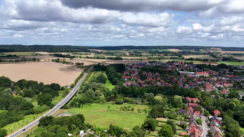 Rural road of historic town in Germany during sunny day with clouds. Agricultural farm fields and hills in distance. Red tile roof houses in small town of America. Aerial wide shot.