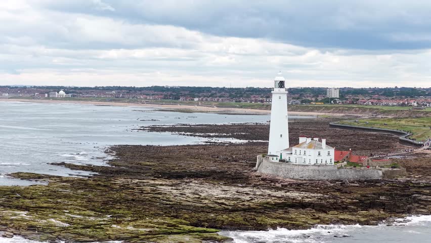 Aerial camera smoothly pans across St Maryâs Lighthouse and surrounding rocky shoreline under overcast daylight, revealing coastal landscape and distant townscape in Whitley Bay, England