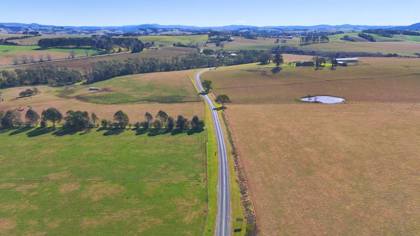 Drone footage glides above a quiet country road, passing open fields, scattered trees, and small ponds under bright daylight in rural New South Wales