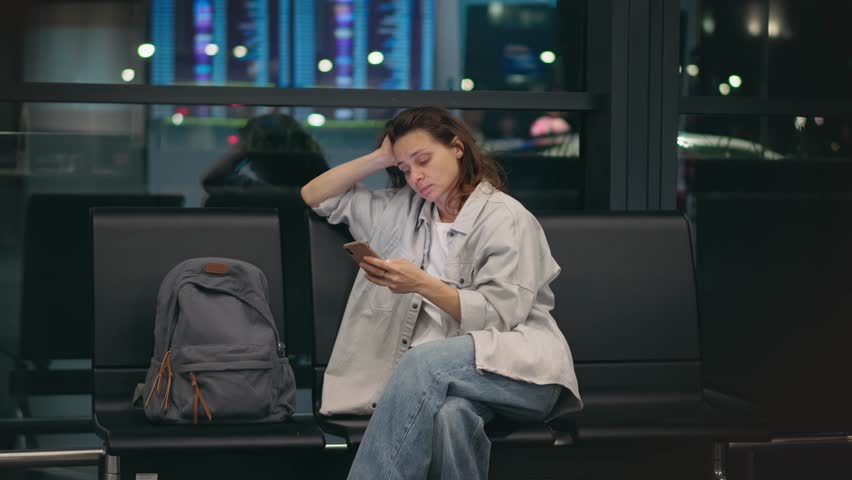 A young, tired woman is sitting at the airport with a phone in her hands, waiting for her night flight