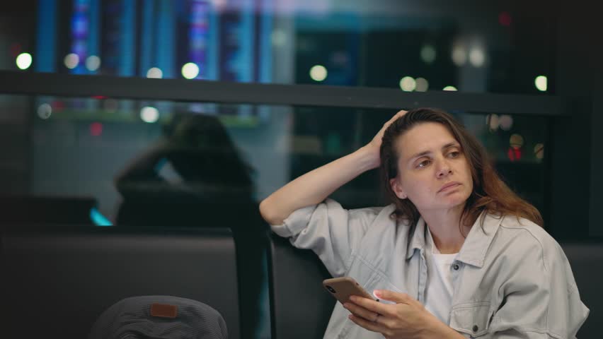 A young, tired woman is sitting at the airport with a phone in her hands, waiting for her night flight