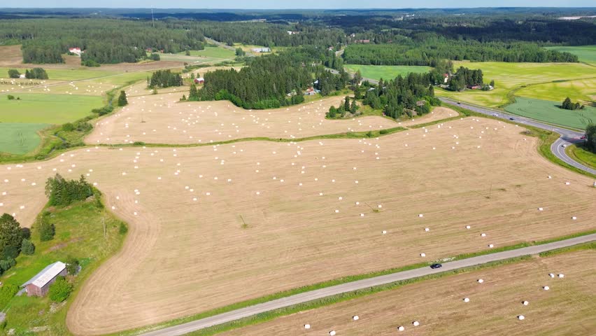 A rural landscape with hay bales scattered across a field. A car drives along a dirt road that cuts through the farmland. A tractor is parked in the field.