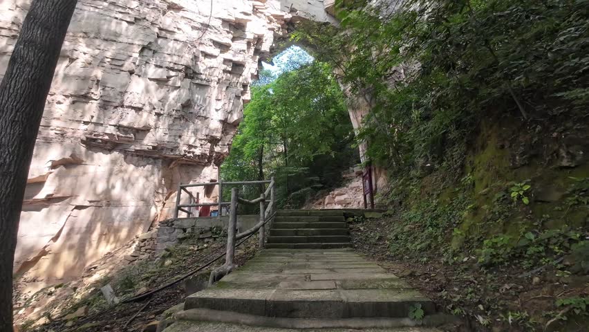 Natural stone arch and stairs surrounded by dense forest, camera rising in Zhangjiajie on a sunny day.