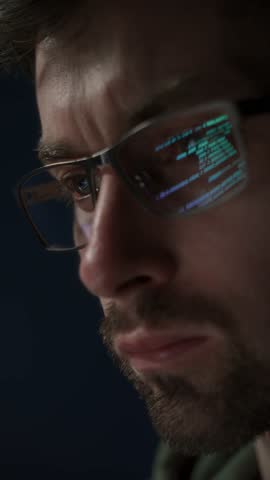 Close-up a focused data scientist wearing glasses, with lines of computer code reflecting on his lenses. Software engineer analysing data late in a dark room. Vertical video