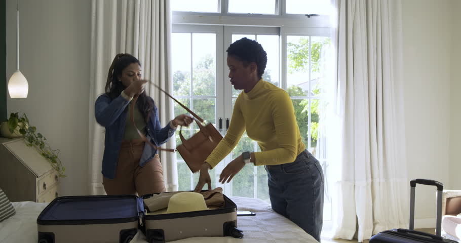 Diverse female friends zipping suitcase on bed in bedroom, pulling smartphone for selfie. Travel, friendship, casual, lifestyle, modern, vibrant, leisure