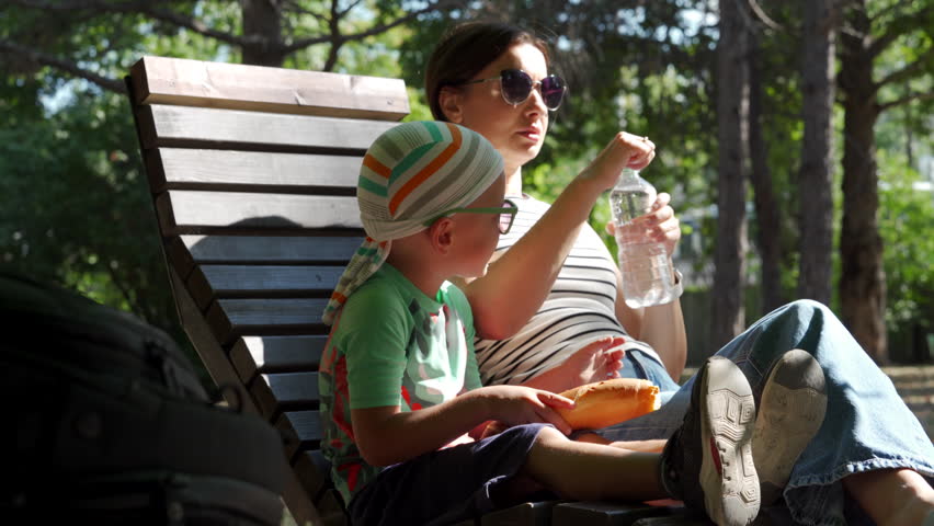 A young boy drinks water while sitting next to his mother on a wooden bench. They relax in a shaded park surrounded by trees on a warm day.