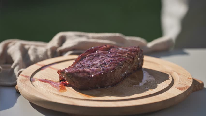 A cook is cutting freshly grilled juicy beef steak on wooden cutting board in backyard garden, food closeup, outdoor cooking