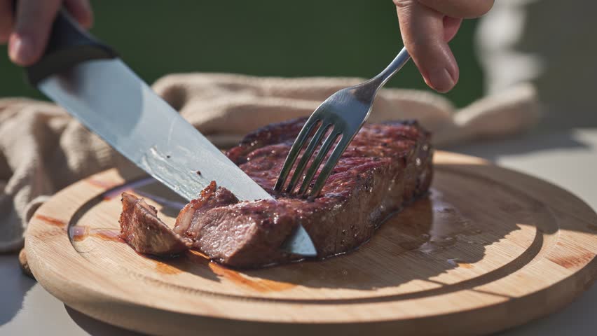 A cook is cutting freshly grilled juicy beef steak on wooden cutting board in backyard garden, food closeup, outdoor cooking