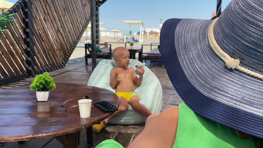 A young child in yellow shorts sits on a light blue bean bag, eating ice cream on a wooden patio. The scene includes a woman in a sunhat watching nearby, with beach umbrellas and sand