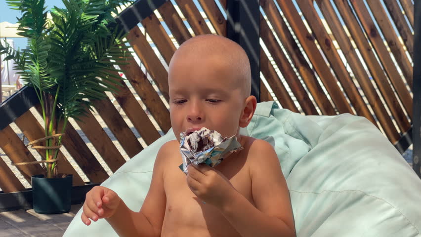 A young child sits comfortably on a light blue bean bag, savoring a chocolate ice cream bar. The toddler is surrounded by a wooden fence and a potted plant, basking in the warm sunlight.