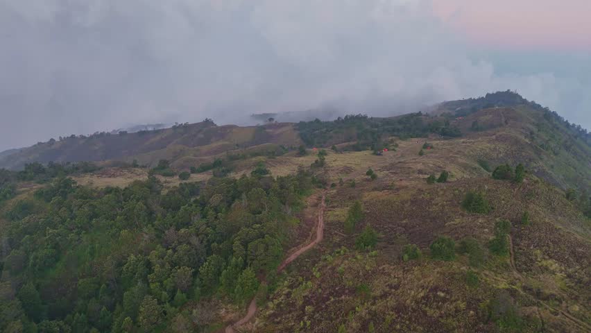 Aerial view of mountain ridge covered in grassland and scattered trees with narrow hiking trail winding through the landscape. Misty clouds roll over the hills in the background. Mount Prau, Indonesia