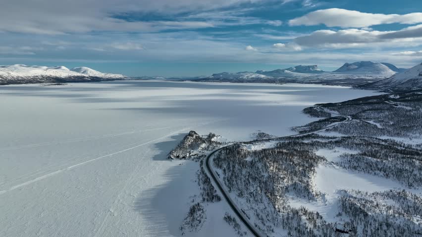 Aerial view of a snowy landscape, with a frozen lake reflecting the sky, a winding road cutting through snow-covered trees, Sweden.