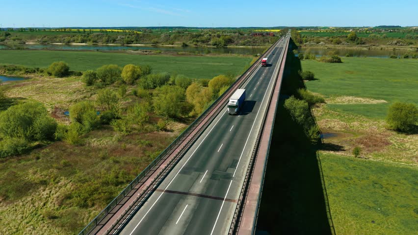Heavy duty truck with cargo trailer driving across bridge through countryside landscape. Freight hauler moving on elevated road supporting logistics and delivery services. Long-haul transport vehicle