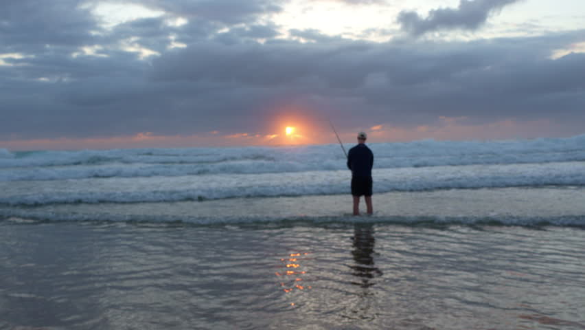 Wide Shot of Man Fishing on Beach at Sunrise – Orange Sun Rising Over Ocean Waves in Early Morning Light