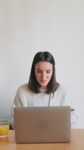 Vertical video, A young woman works from her home office, enjoying a glass of orange juice while multitasking on her laptop and phone.