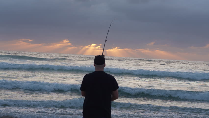 Man Fishing at Dawn on Beach – medium shot from behind with waves rolling In under soft morning light and casting a line into the sea as soft waves lap the shore, captured from behind