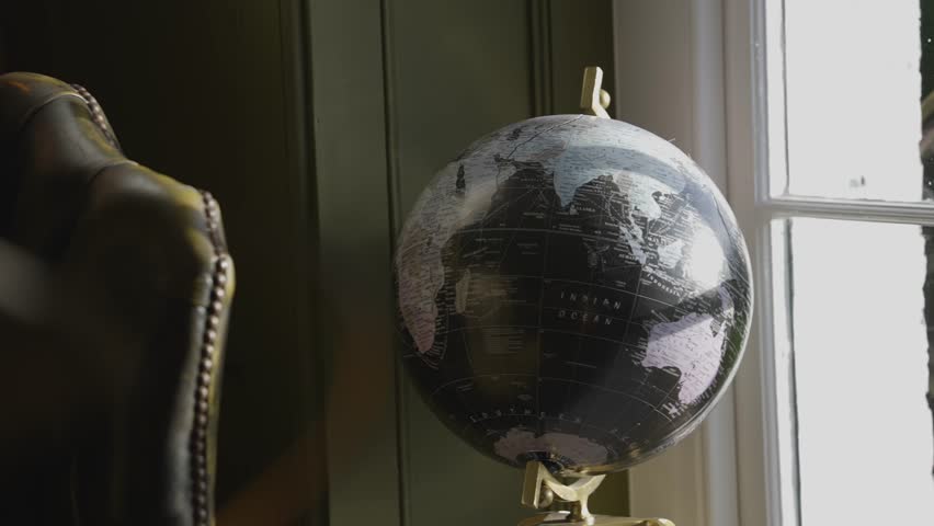 A globe sits on a stand in front of a window. The globe is black and white and has a map of the world on it. The room is dimly lit, giving the scene a cozy and intimate atmosphere