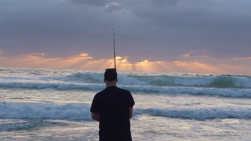 Medium shot of man fishing on beach at dawn as sun rays break through clouds. Waves crashing on breach as he casts and reels in fishing line. 