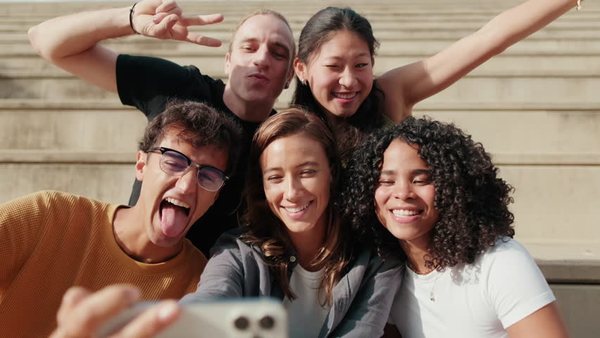 Happy multiethnic group of university colleagues sitting on stairs outdoors in the city, using smartphone to take a mobile photo, laughing and having fun together. Technology addicted