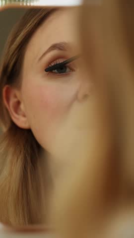 A woman focuses on her reflection while applying eyeliner. The setting is a well-lit bathroom, highlighting her careful makeup application as part of her daily routine.