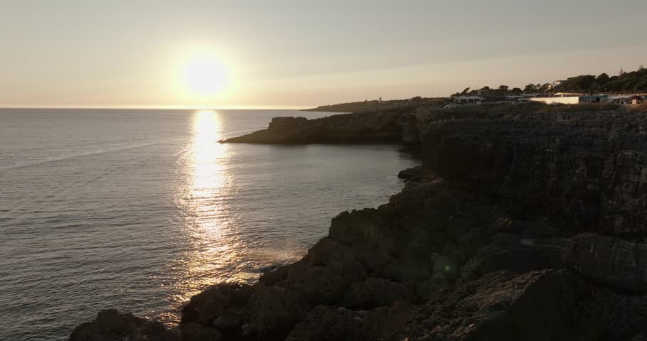 Aerial view of the sun setting over the rocky cliffs and calm sea along the Algarve coastline in Portugal.