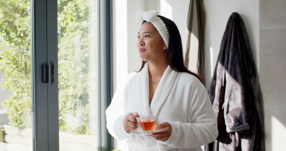 Asian woman entering bathroom holding teacup and saucer gazing at doors sipping tea savoring calm. Serenity, tranquility, mindfulness, reflection, wellness, natural light, minimalist