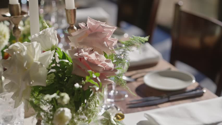 Parallax shot focusing on floral arrangement atop a wedding table, with blurred napkins, candles, and cutlery in the background, creating a dreamy and elegant dining atmosphere.