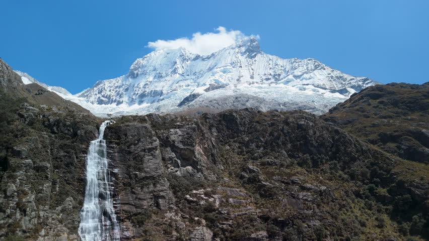 Stunning aerial shot of a glacier-fed waterfall plunging from Chacraraju mountain, feeding Laguna 69 and surrounded by the dramatic peaks of the Cordillera Blanca in Peru.