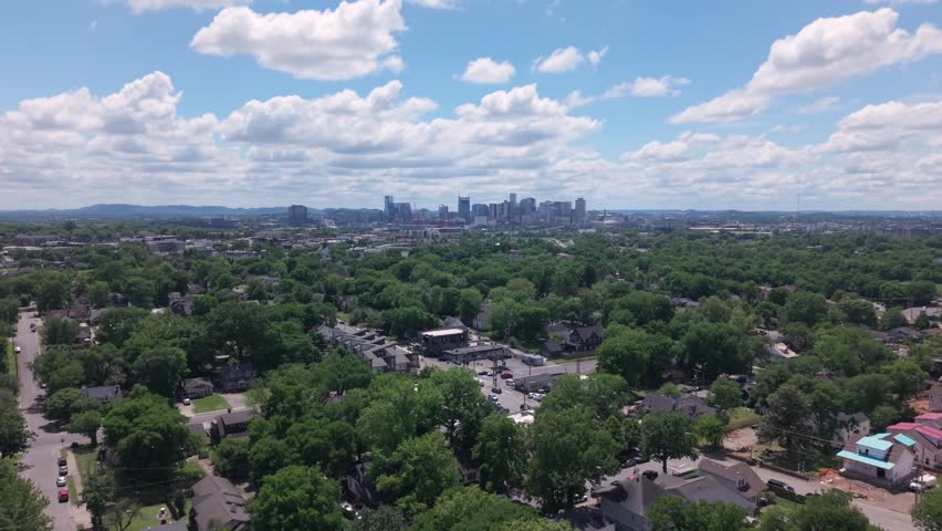 Lush green trees covering residential area with Nashville skyline in background under beautiful cloudy sky. Aerial, establishing push forward